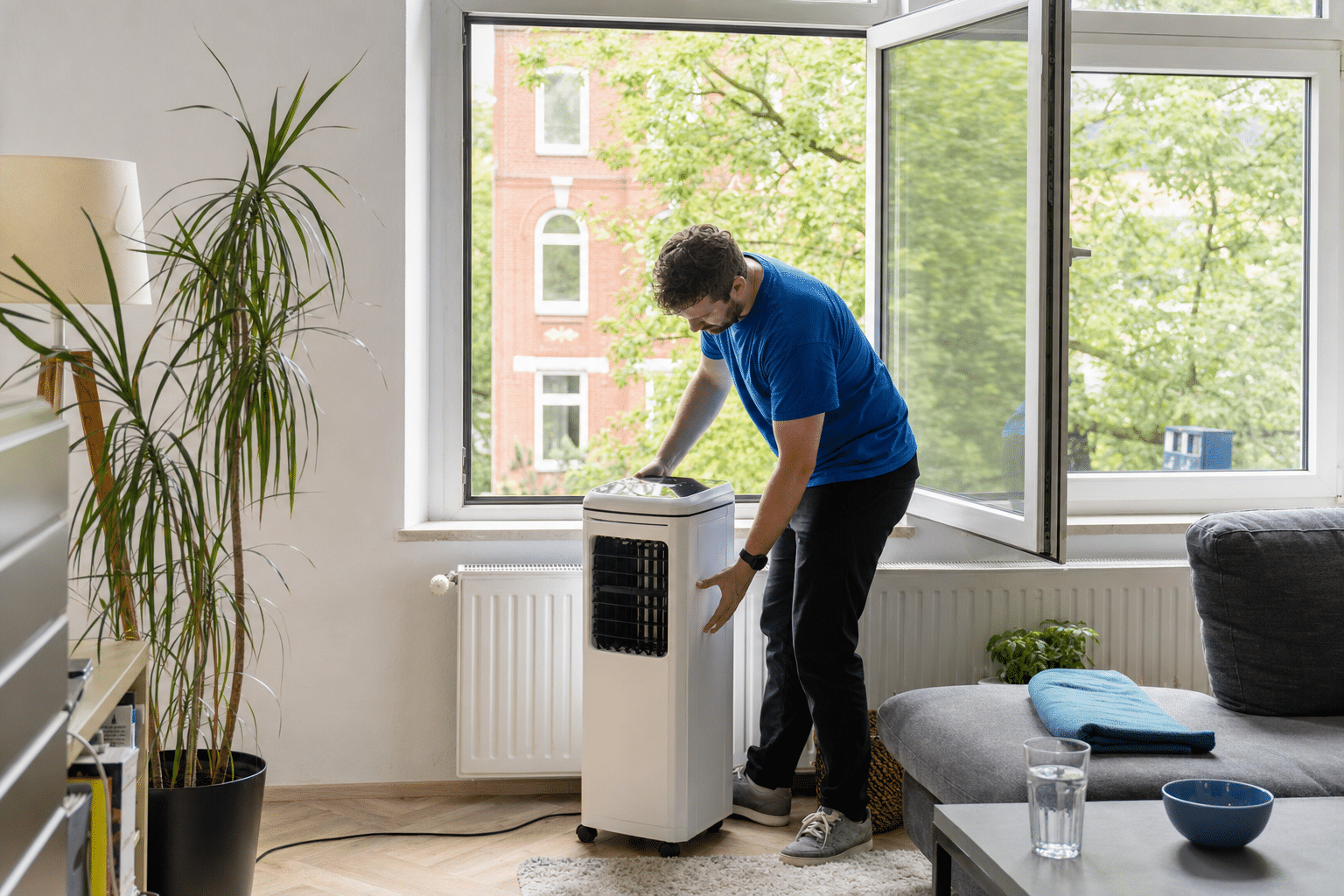Eine Person stellt ein mobiles Luftkühlgerät neben ein geöffnetes Fenster im Wohnzimmer an einem heißen Sommertag